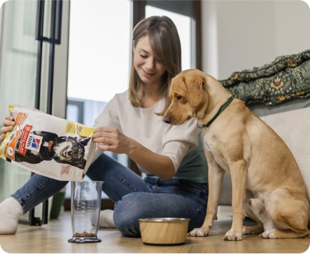 Une femme verse de la nourriture Hill's Pet Nutrition dans un récipient gradué tandis qu'un chien l'observe attentivement.